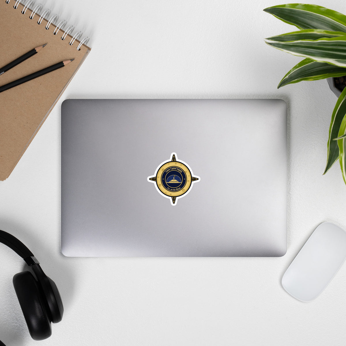3 inch Lunar Capitol Dome Crest Sticker on a closed laptop with headphones, notebook and a plant