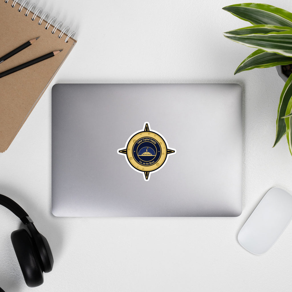 4 inch Lunar Capitol Dome Crest Sticker on a closed laptop with headphones, notebook and a plant