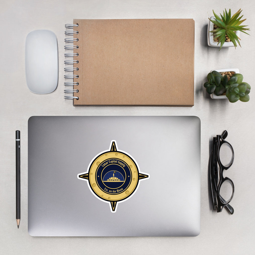 5.5 inch Lunar Capitol Dome Crest Sticker on a silver laptop lid beside a kraft notebook, mouse and glasses