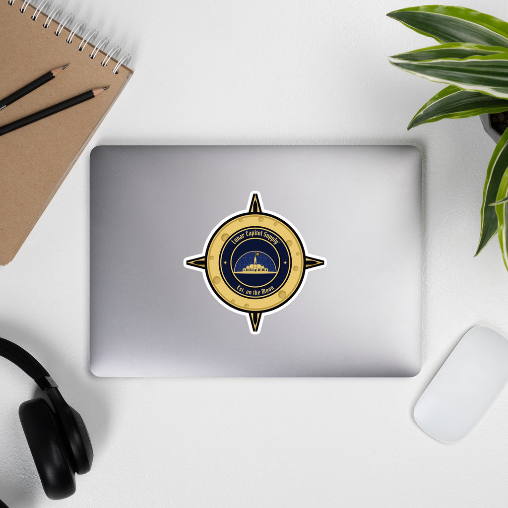 5.5 inch Lunar Capitol Dome Crest Sticker on a closed laptop with headphones, notebook and a plant
